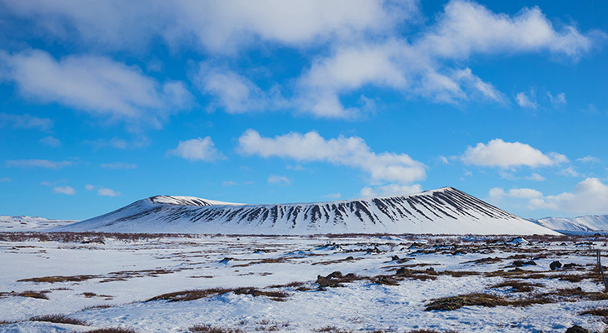 Hverfjall-krater Besneeuwd berglandschap met donkere lavastrepen onder een blauwe lucht met wolken.