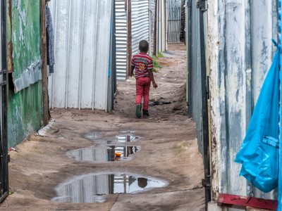 Jongen loopt door smalle steeg met golfplaten huizen en plassen water in Zuid-Afrika.