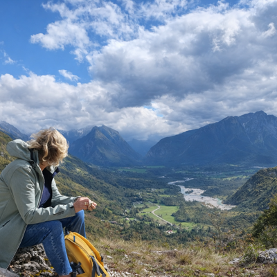 Wandelaar met rugzak kijkt uit over een bergvallei in Slovenië, met rivier en bergen onder een dramatische lucht.