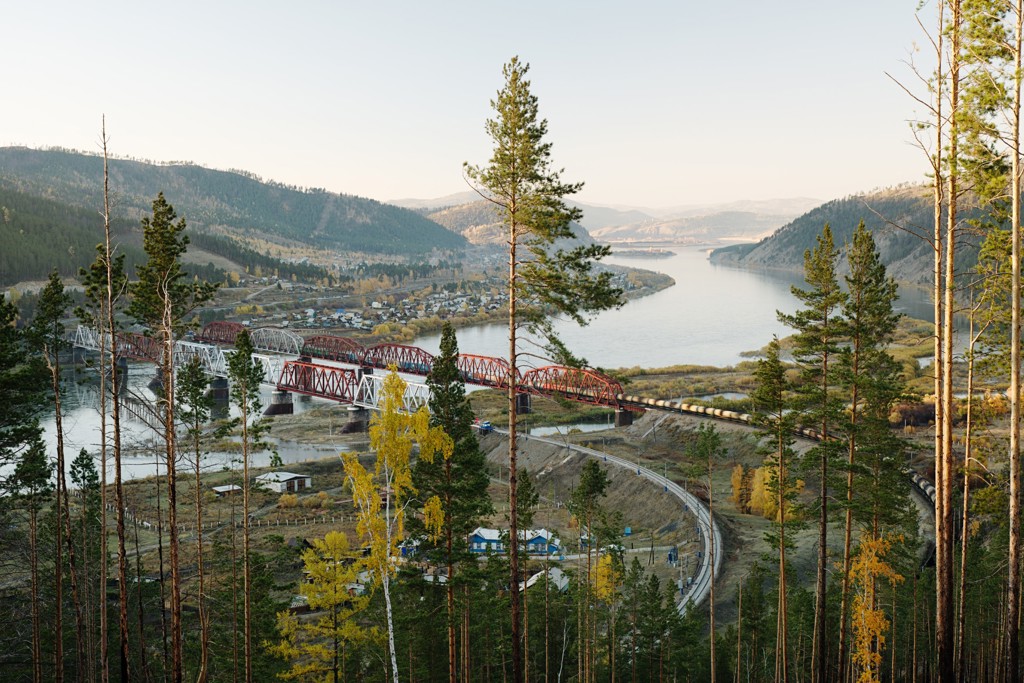 Uitzicht op de Trans-Siberië spoorlijn met trein over een rode brug langs een rivier, tussen bergen en dennenbos.