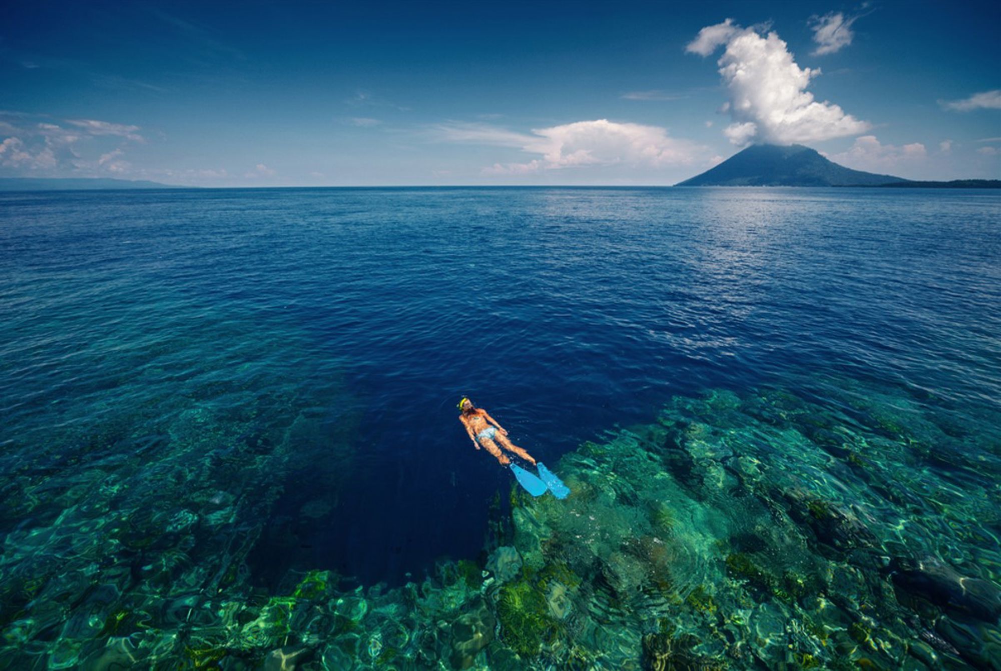 Snorkelaar boven koraalrif in helderblauw water, met vulkaaneiland op de achtergrond in Indonesië.