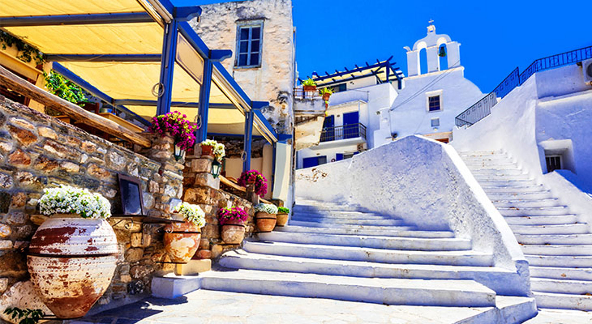 Mykonos Witte trap in Grieks eilanddorp met blauwe luiken, kerk en bloempotten onder een zonnige hemel.