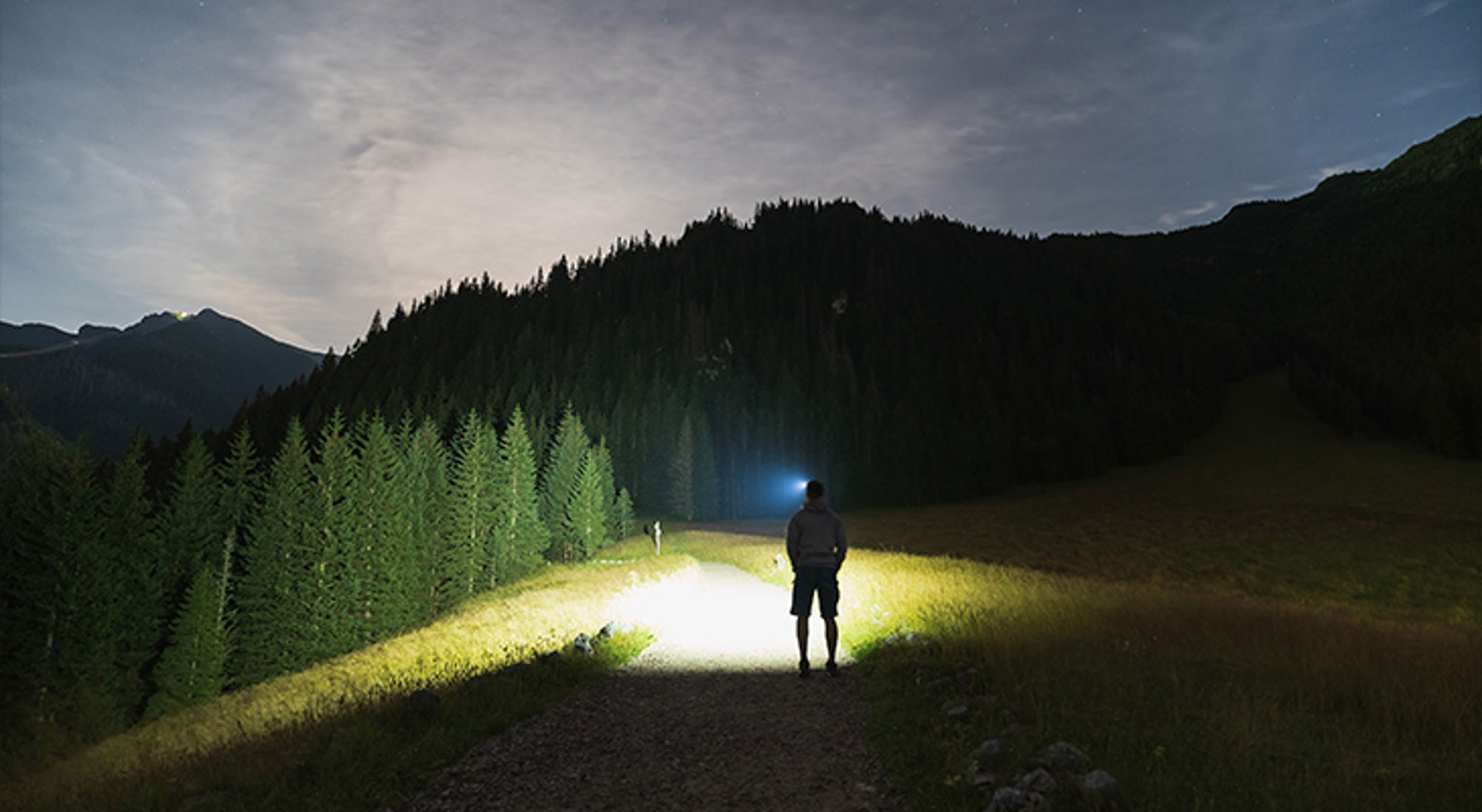 Hoofdlamp Wandelaar met hoofdlamp op bergpad bij nacht, met verlichte bomen en silhouetten van bergen op de achtergrond.