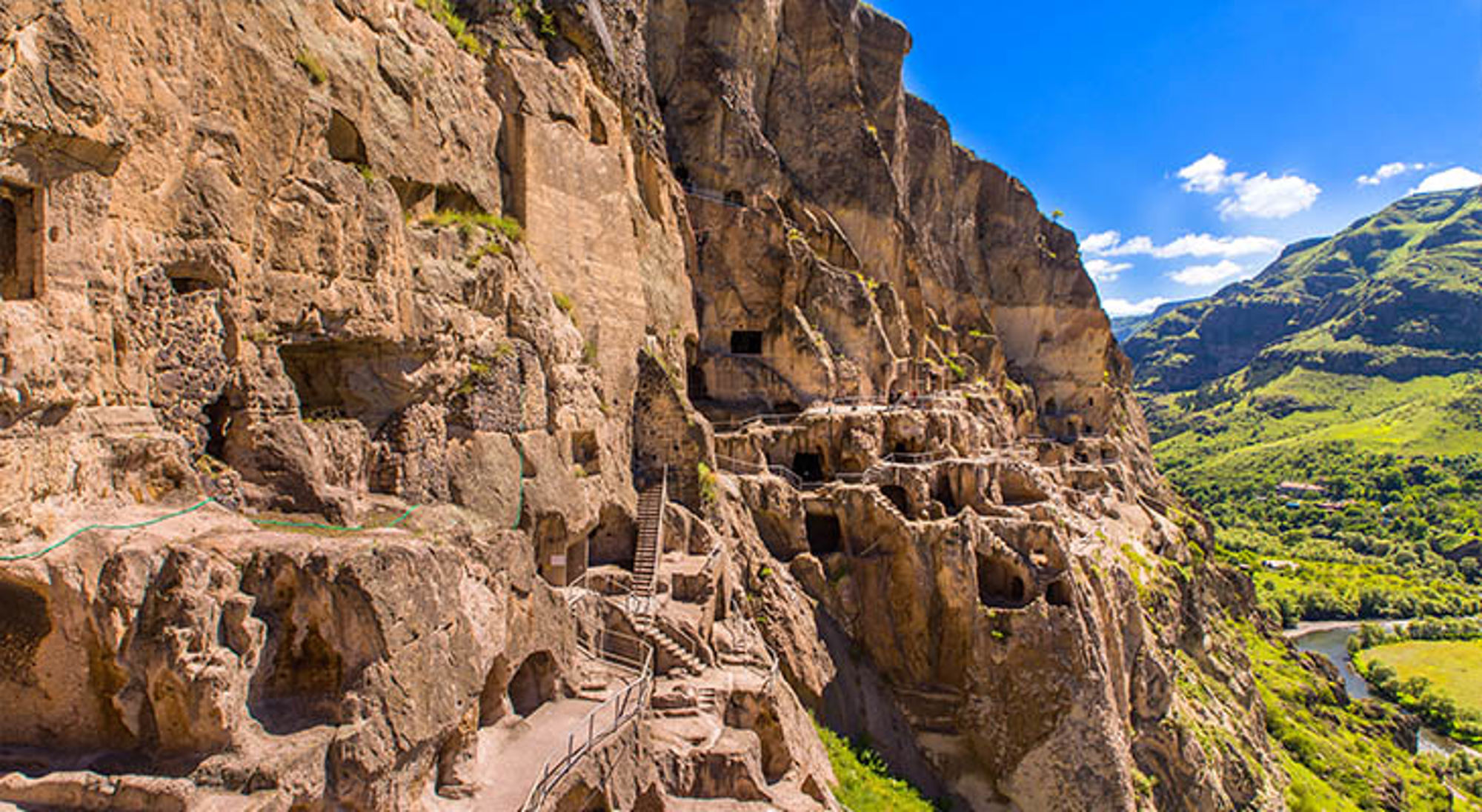 Rotsklooster Vardzia met uitgehakte grotwoningen en wandelpad langs de klif, met groene vallei op de achtergrond.
