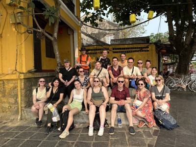 Groepsfoto van reizigers tijdens de Route Vietnam, zittend voor een geel gebouw met lampionnen.