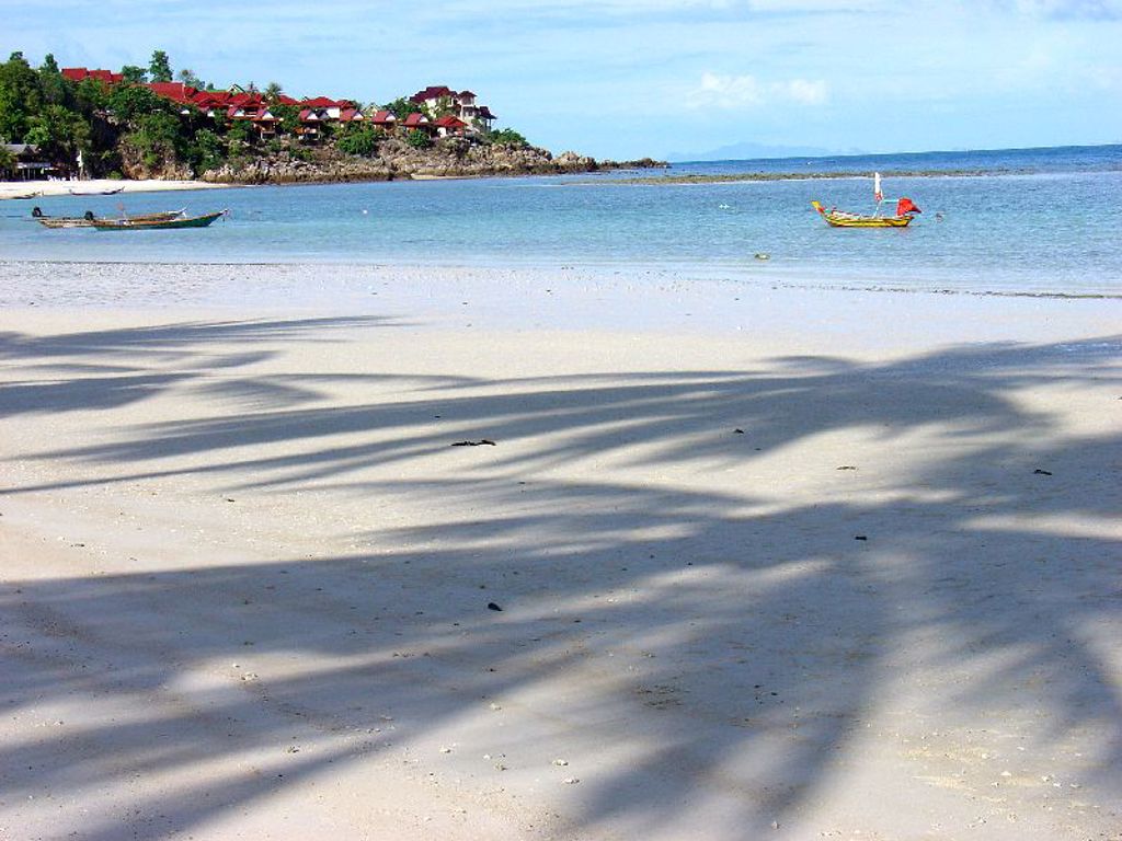 Strand in Thailand met longtailboten op helderblauw water en resort op een groene heuvel.