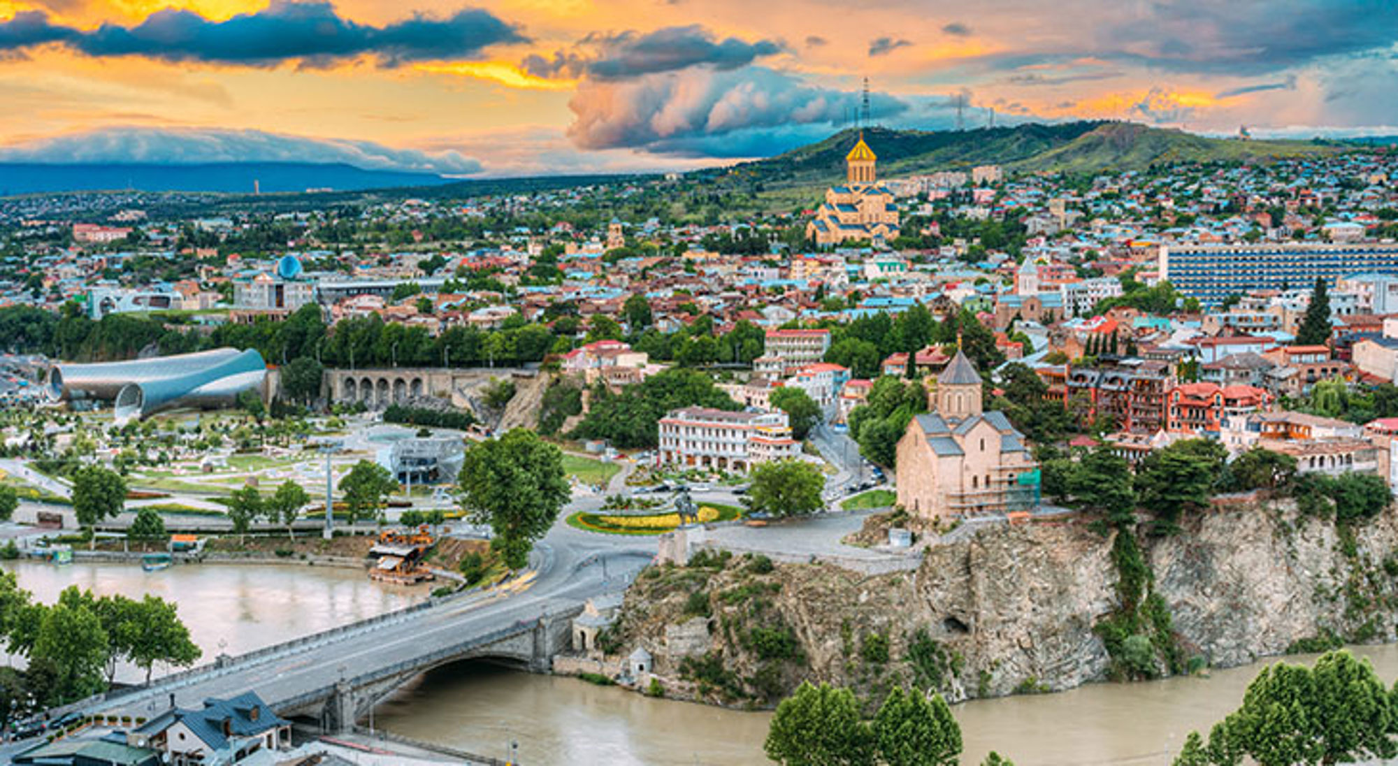 Panorama van Tbilisi met de Metekhi-kerk aan de rivier en de Heilige Drie-eenheidskathedraal op de heuvel bij zonsondergang.