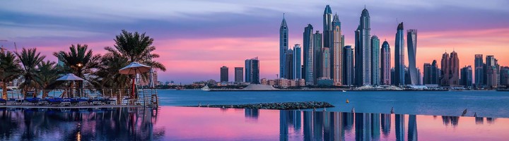 Skyline van Dubai bij zonsondergang met moderne wolkenkrabbers aan het water en palmbomen op de voorgrond.