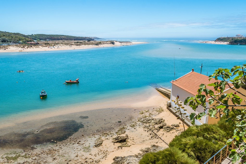 Uitzicht op turquoise riviermonding en strand aan de Costa Vicentina in Portugal, met vissersbootjes en wit huis.