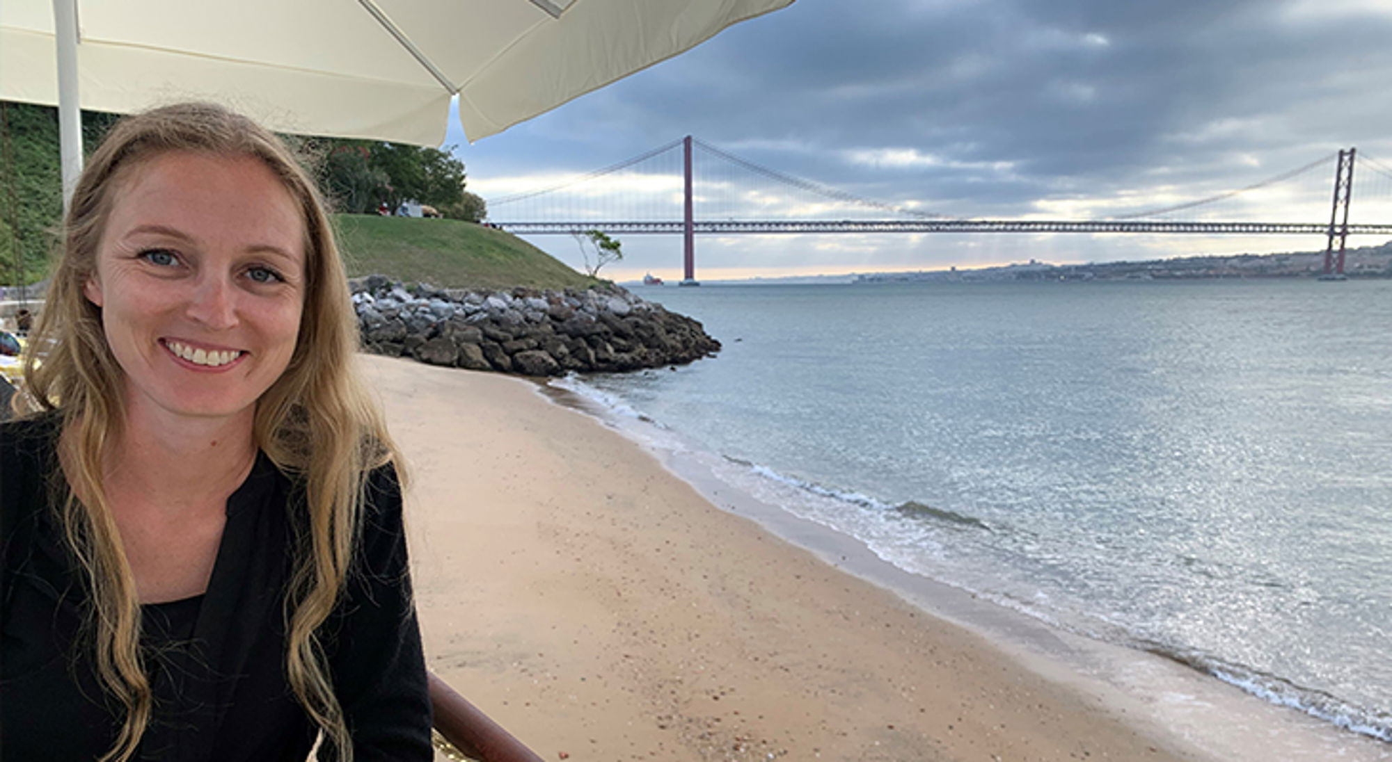 Monique Vrouw op een terras aan het strand met de Ponte 25 de Abril-brug op de achtergrond in Lissabon.