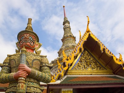 Gouden tempelwachter bij Wat Phra Kaew in Bangkok, met kleurrijk tempeldak en pagode op de achtergrond.