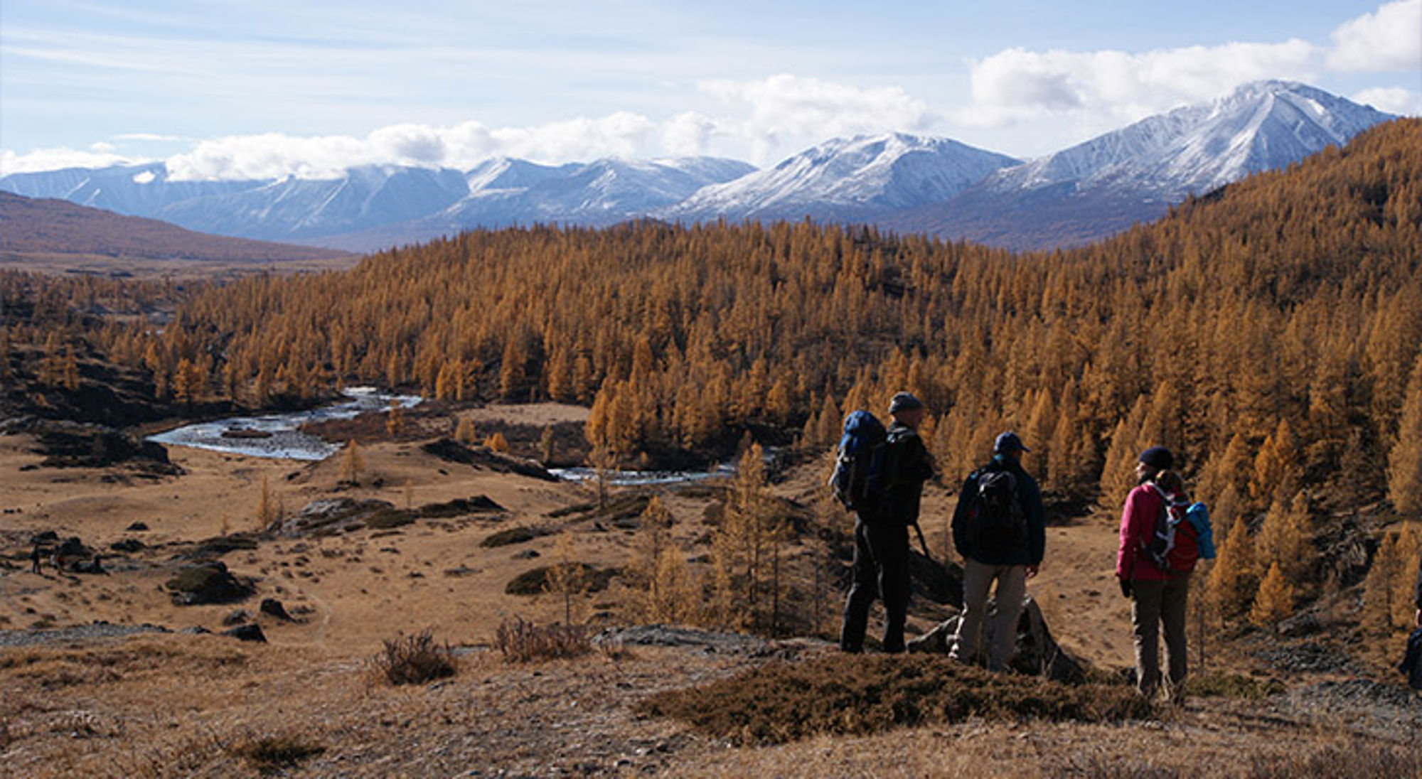 Mongolië Wandelaars met rugzakken kijken uit over een vallei met rivier, herfstbos en besneeuwde bergen.