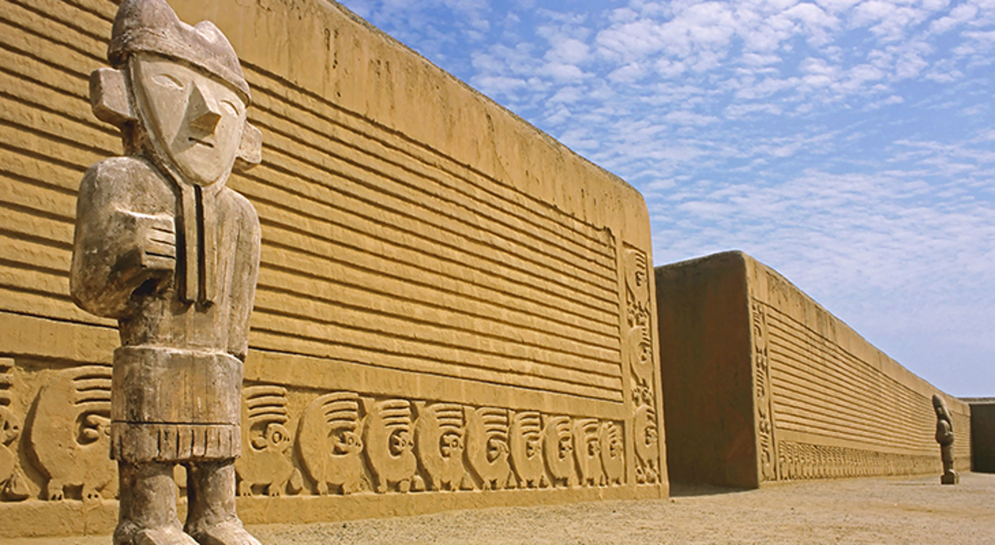 Houten beeld bij de Chan Chan-ruïnes met reliëfmuren en blauwe lucht in Peru.
