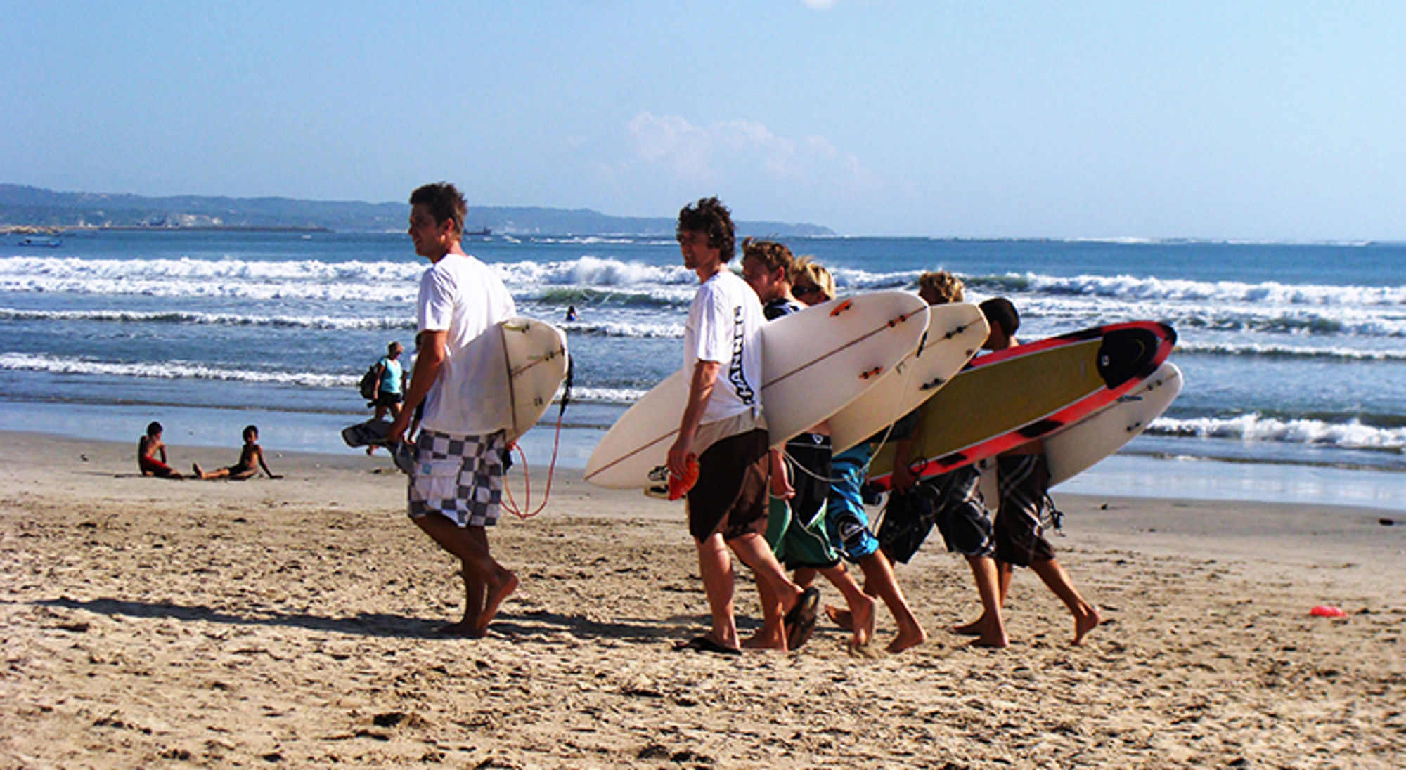 Surfers lopen met surfboards over het strand richting de zee op een zonnige dag.