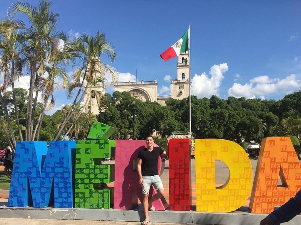 Reiziger bij het kleurrijke MERIDA-bord in Mérida, Mexico, met Mexicaanse vlag en kathedraal op de achtergrond.