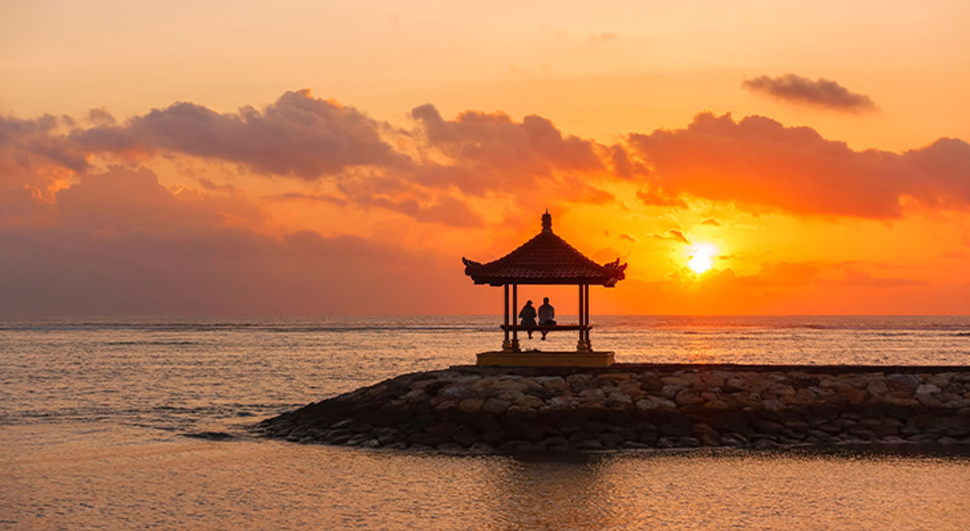Bali, Indonesië Zonsondergang boven zee met silhouet van een prieel op een rotsdam en twee personen die uitkijken.