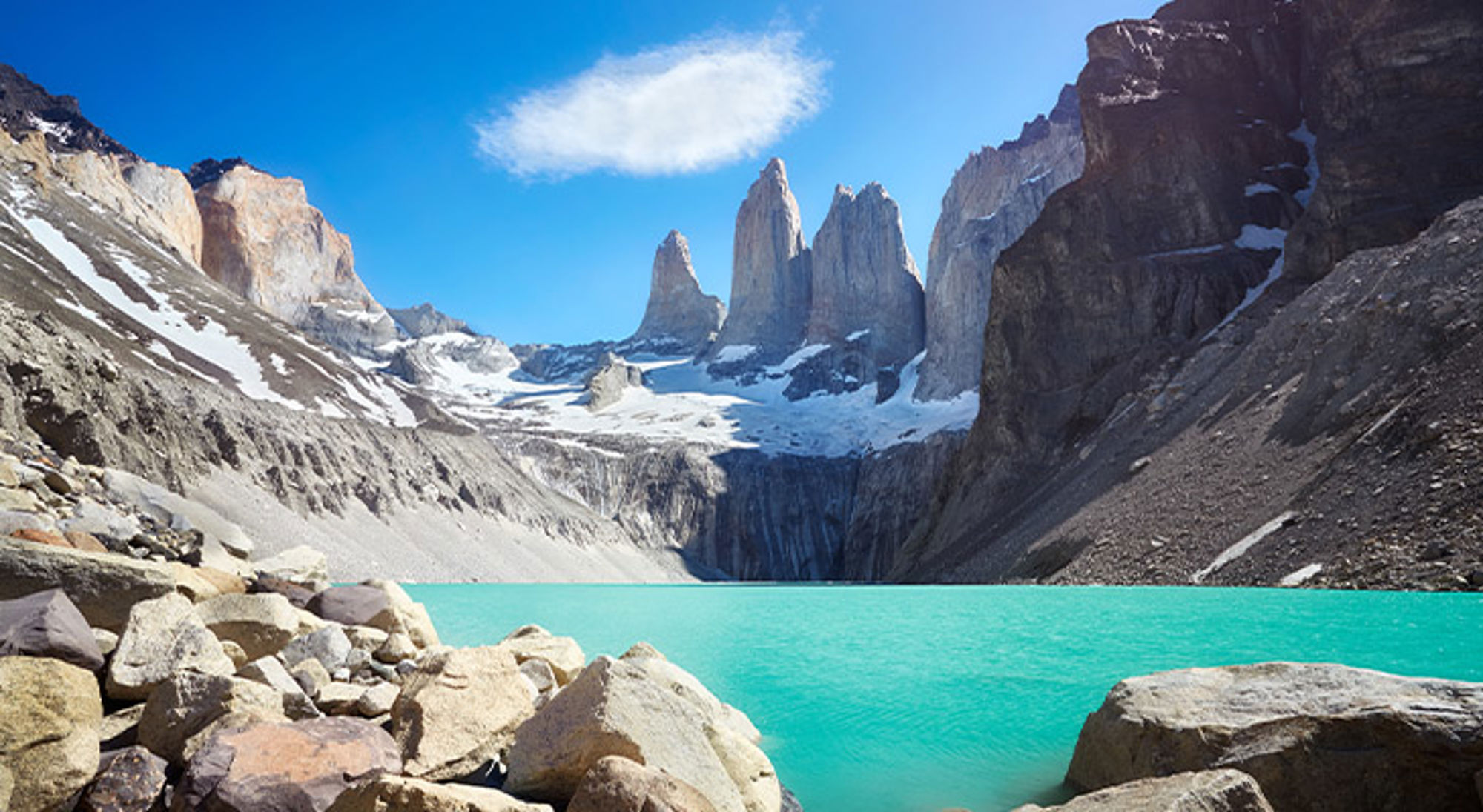 Torres del Paine Turquoise bergmeer met rotsen op de voorgrond en hoge granieten pieken met sneeuw onder een heldere blauwe lucht.
