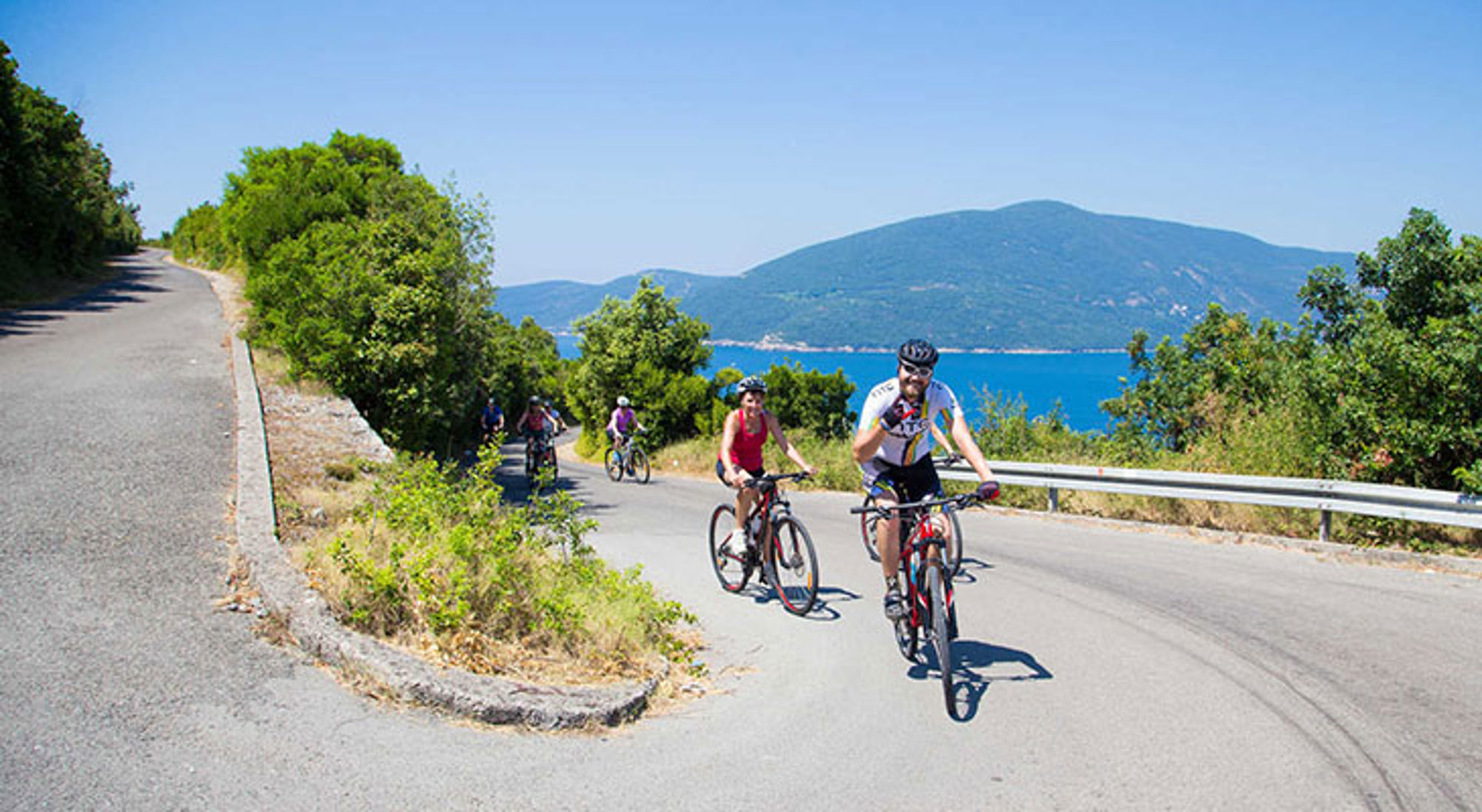 Fietsers op een heuvelachtige kustweg met uitzicht op zee en bergen onder een heldere blauwe lucht.