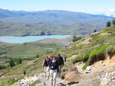 Wandelaars op bergpad met uitzicht op azuurblauw meer en heuvels in Patagonië.