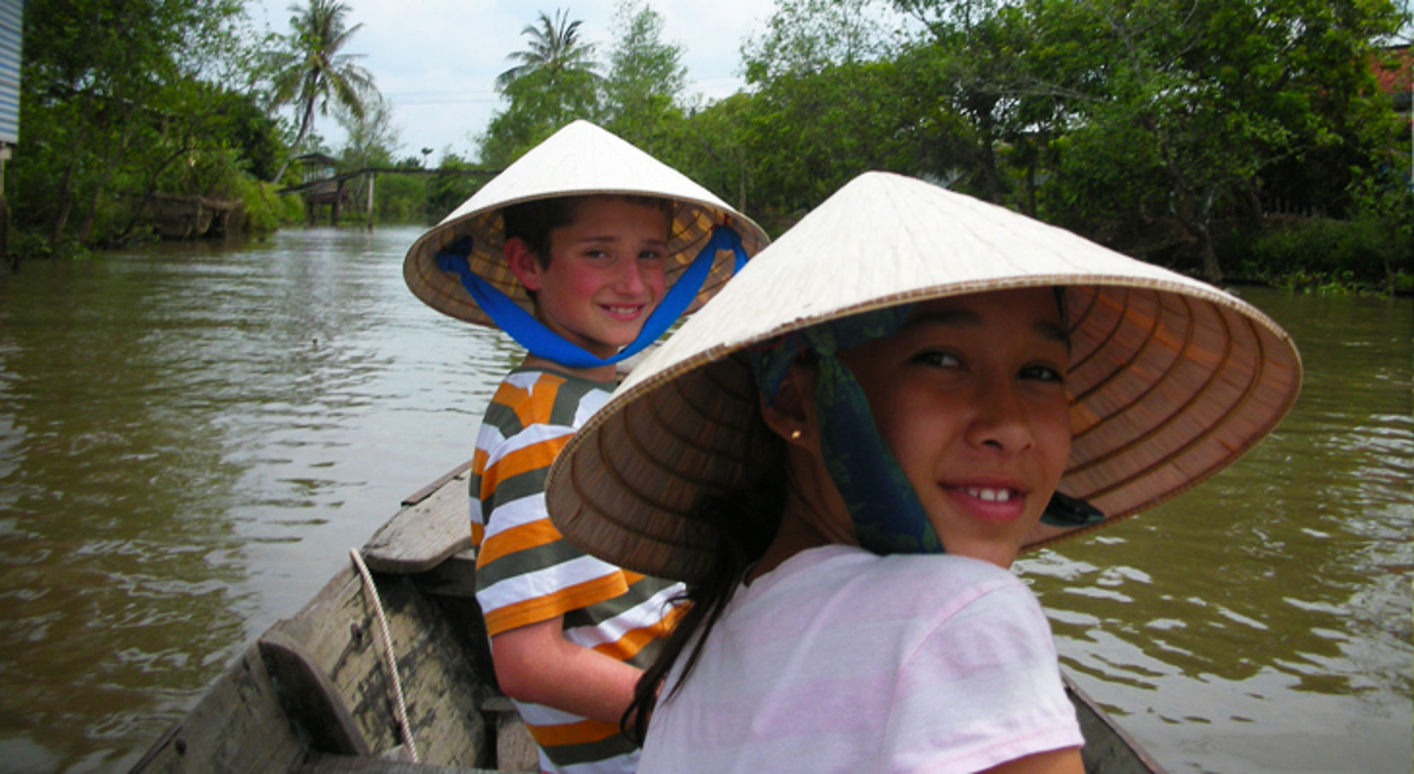 Twee kinderen met Vietnamese hoedjes in een boot op een rivier, met groene oevers op de achtergrond.