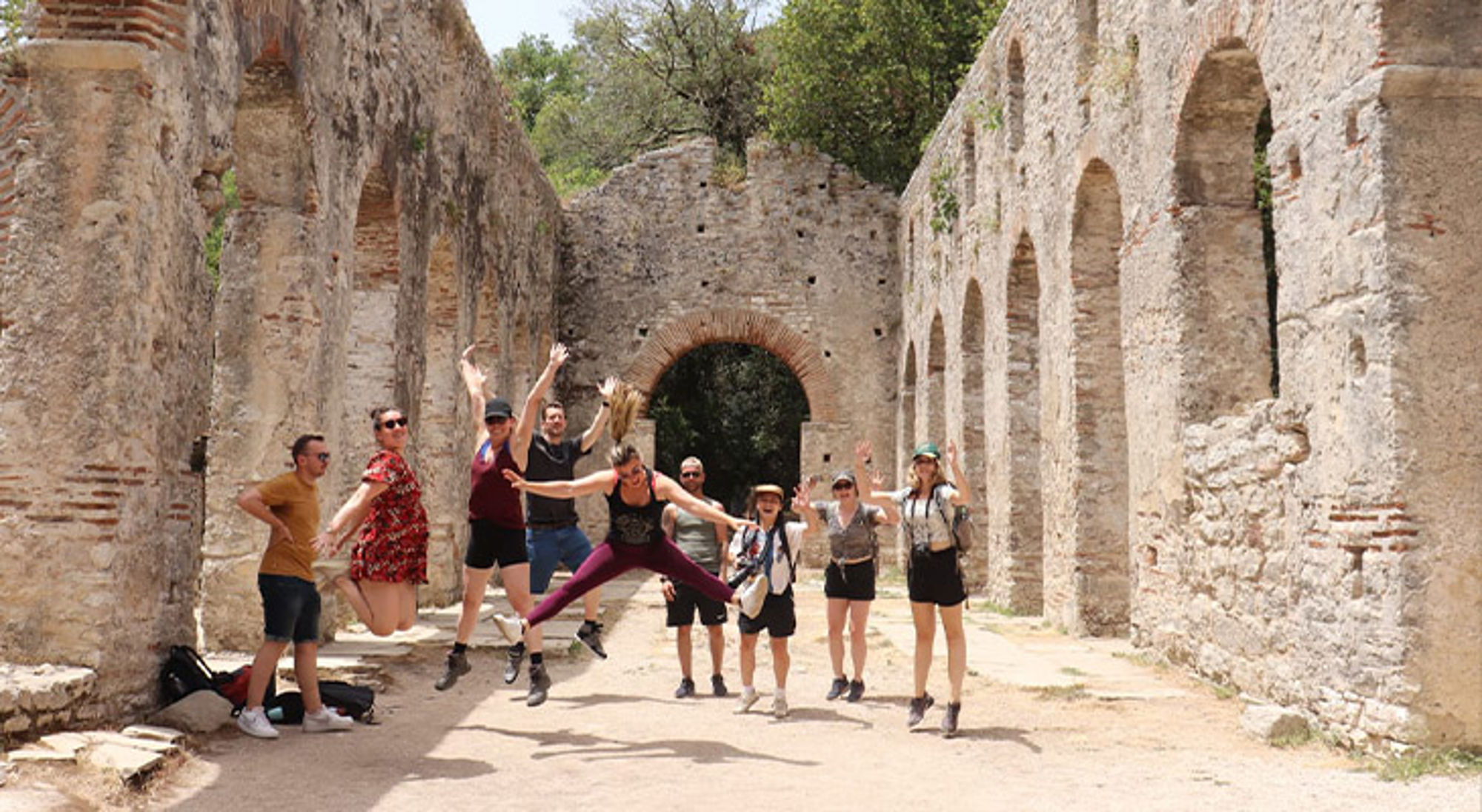 Butrint Albanië Groep reizigers springt en poseert in een oude ruïne met bogen tijdens een excursie.