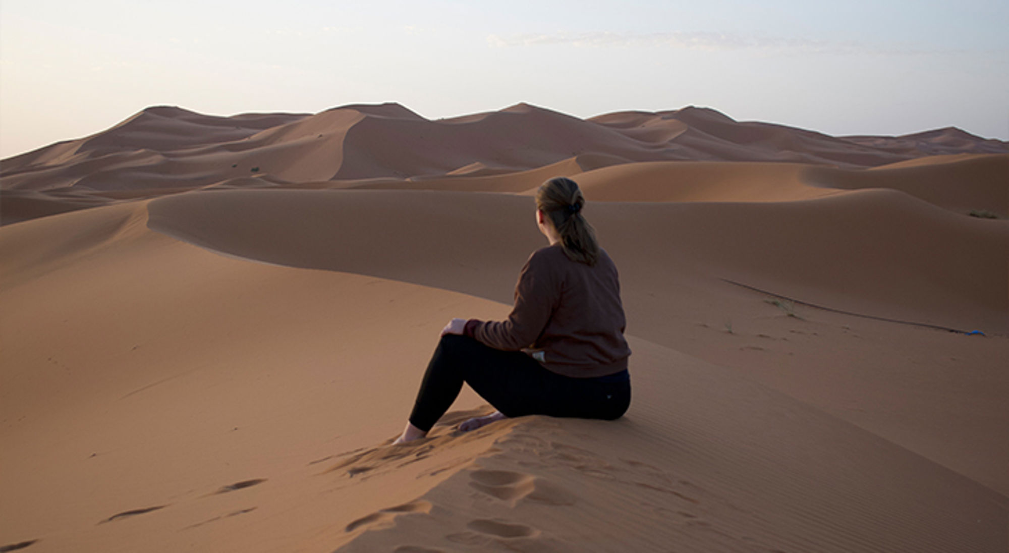 Carolien in de woestijn Vrouw zit op een zandduin en kijkt uit over uitgestrekte woestijnduinen bij zonsondergang.