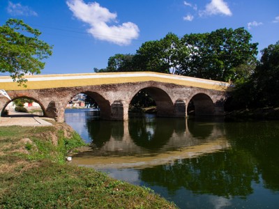 Oude stenen boogbrug over een rustige rivier in Cuba, met bomen en blauwe lucht.