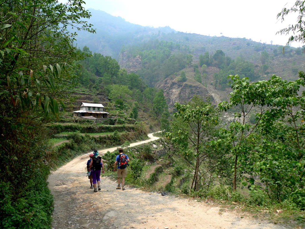Twee wandelaars met rugzakken op een bergpad in Nepal, met groene heuvels en een huisje op de achtergrond.