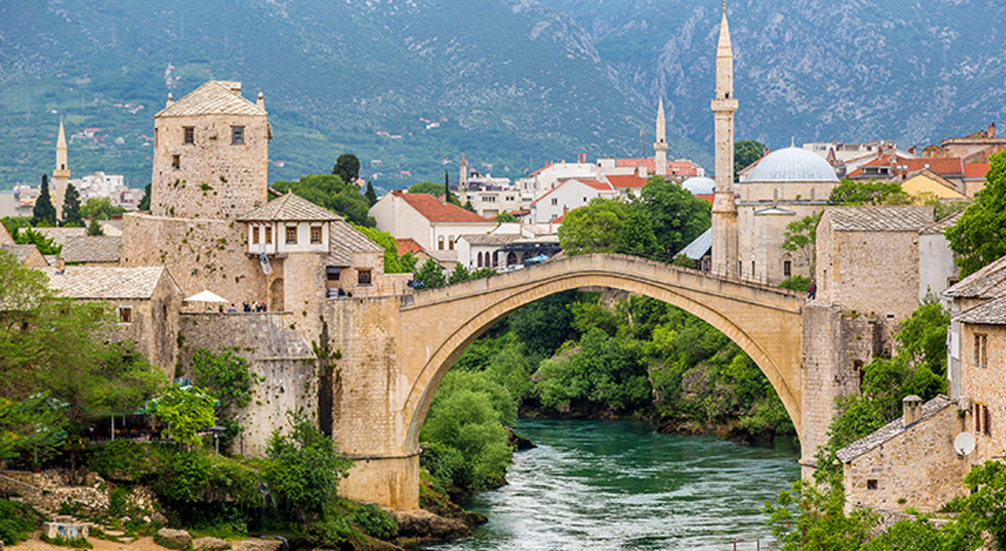 Stari Most-brug over de rivier Neretva in Mostar, met historische gebouwen en minaretten op de achtergrond.