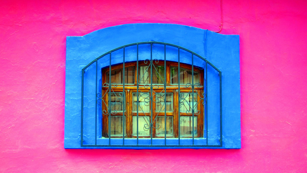 Blauw omlijst raam met smeedijzeren tralies in felroze gevel tijdens groepsrondreis Mexico, Guatemala en Honduras.