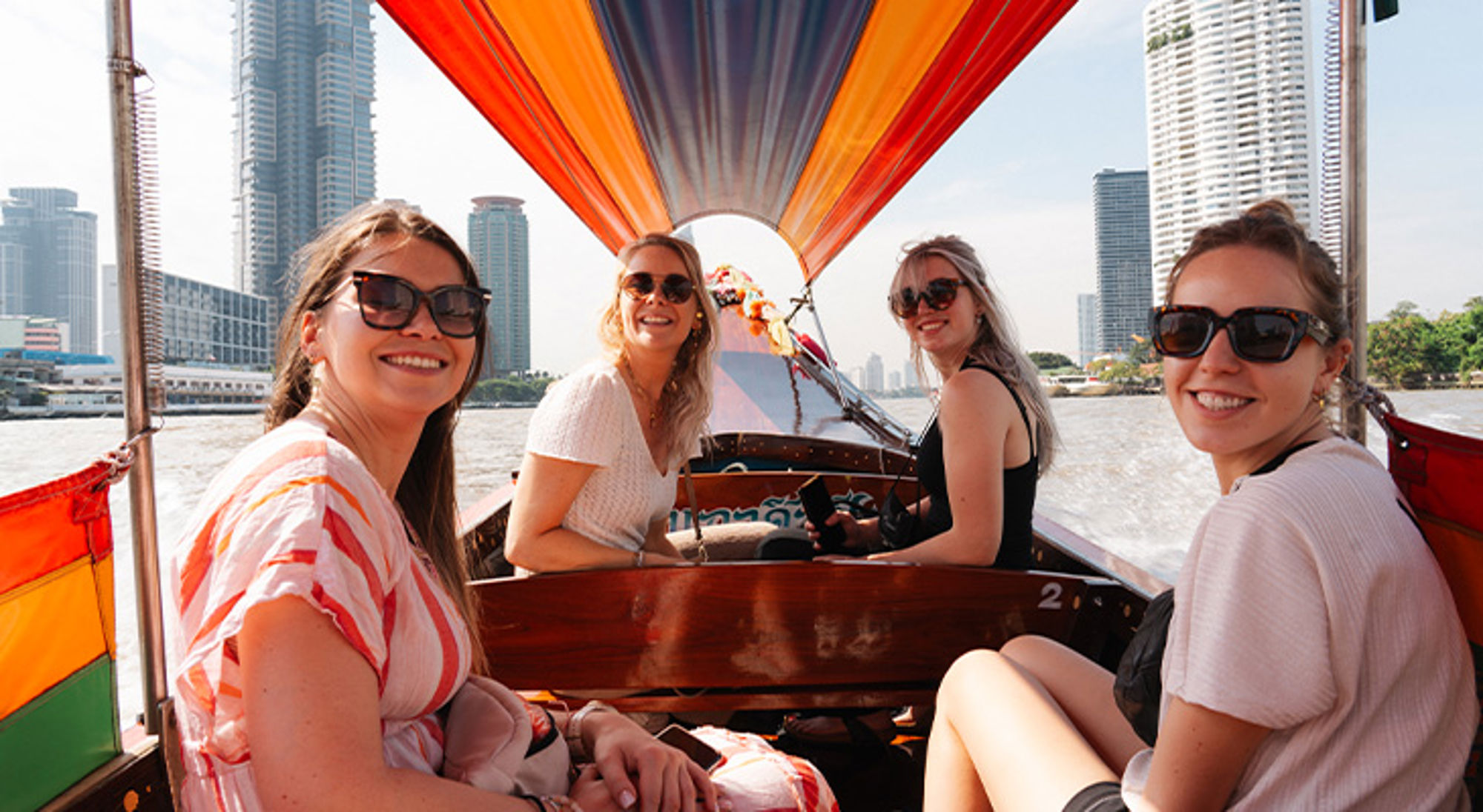 Vier vrouwen in een longtailboot op de rivier, met wolkenkrabbers op de achtergrond.