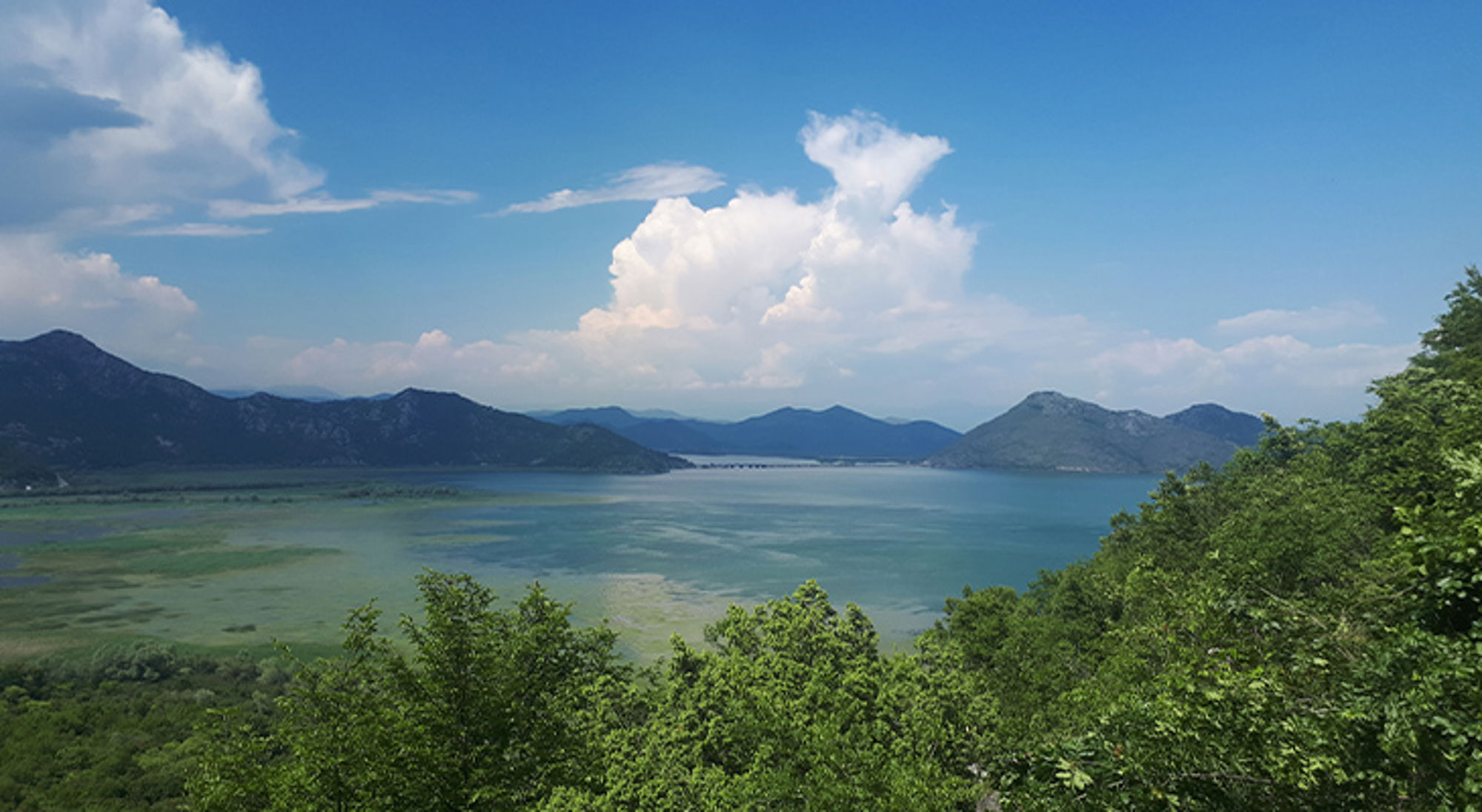 Balkan Panoramisch uitzicht over een meer met omliggende bergen en groene bossen onder een blauwe lucht met wolken.