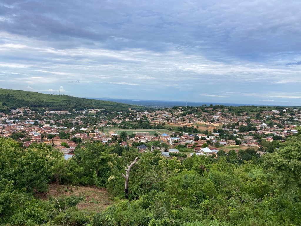 Panoramisch uitzicht over een stad in Ghana, omringd door groen en heuvels onder een bewolkte lucht.