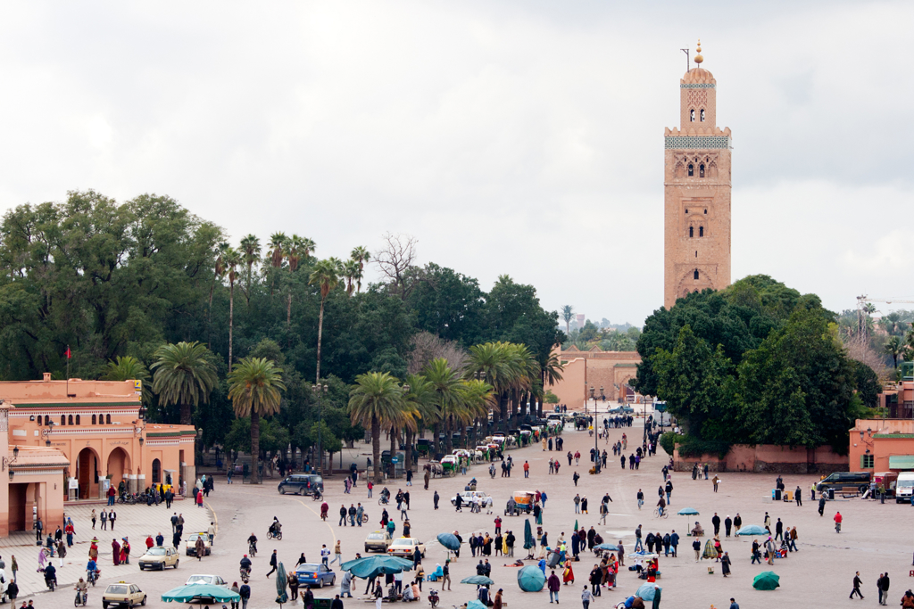 Drukte op het Djemaa el Fna-plein in Marrakech met palmbomen en de minaret van de Koutoubia-moskee.
