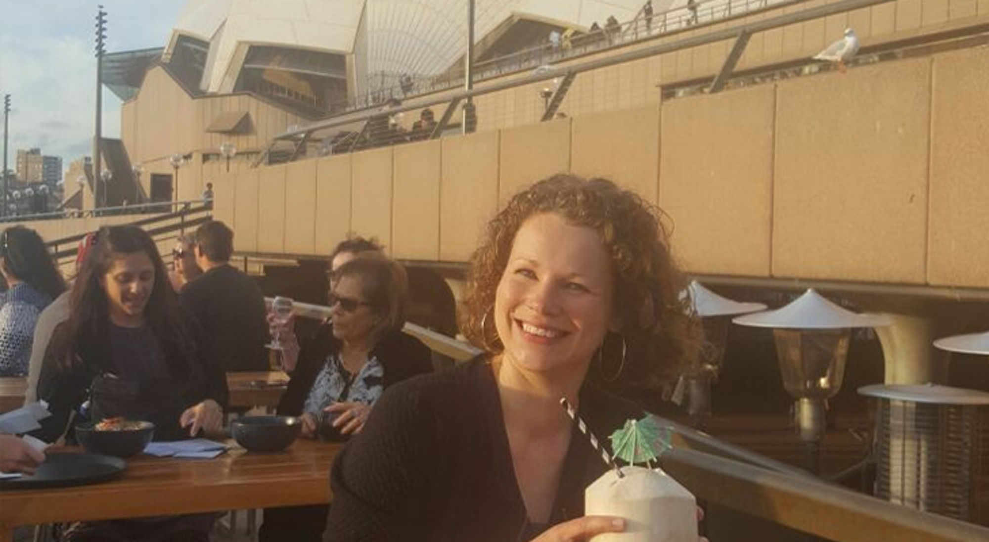 Wendy in Australië Vrouw op dakterras in Sydney met kokosnootdrankje, met het Sydney Opera House op de achtergrond.