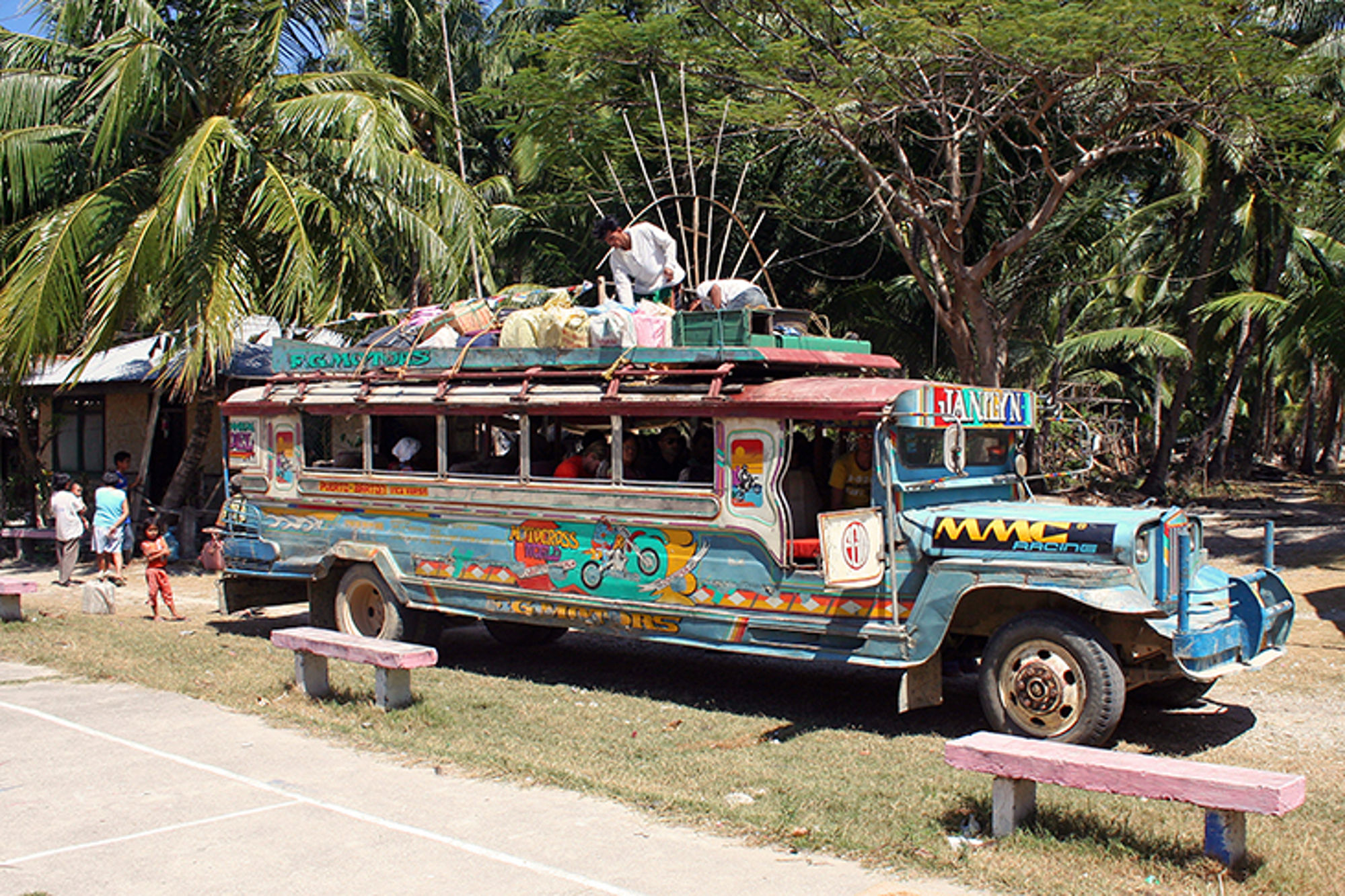 Kleurrijke jeepney met passagiers en bagage op het dak, geparkeerd langs een weg met palmbomen.