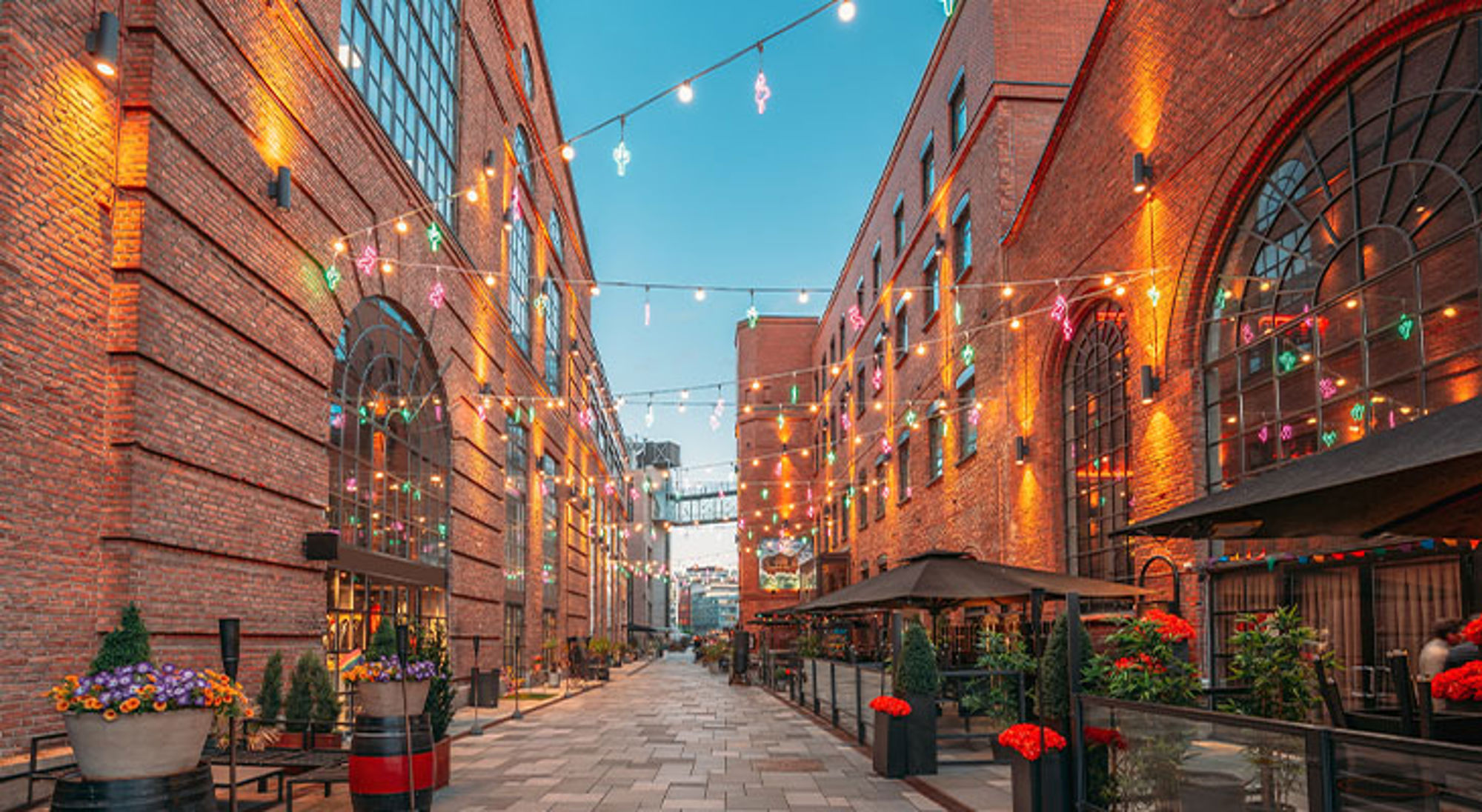 Aker Brygge in Oslo, Noorwegen Verlichte straat tussen bakstenen pakhuizen met terras en slingers van lampjes in de avondschemering.