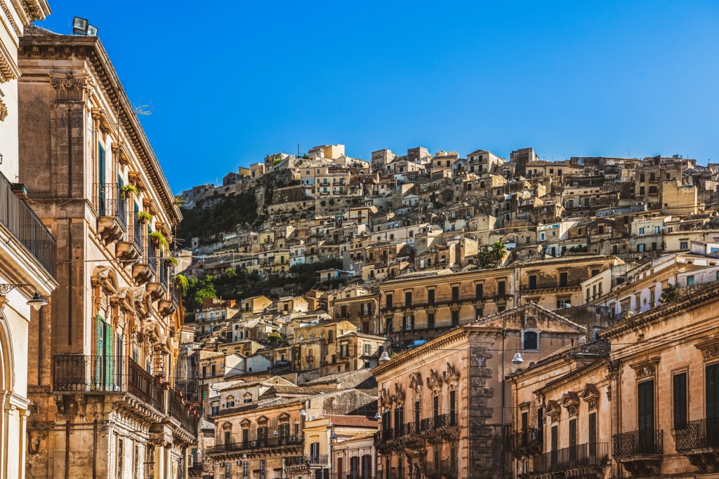 Historische stad in Sicilië met huizen op een heuvel en barokke gevels onder een strakblauwe lucht.