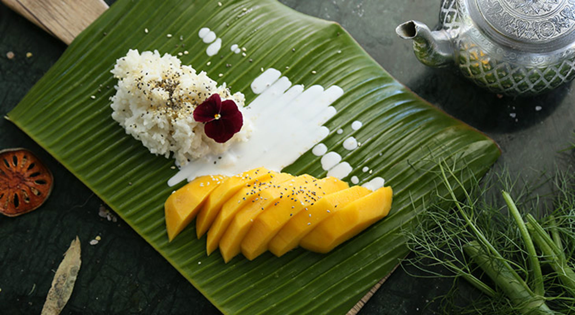 Mango sticky rice met kokosmelk geserveerd op bananenblad, met sesam en eetbare bloem.