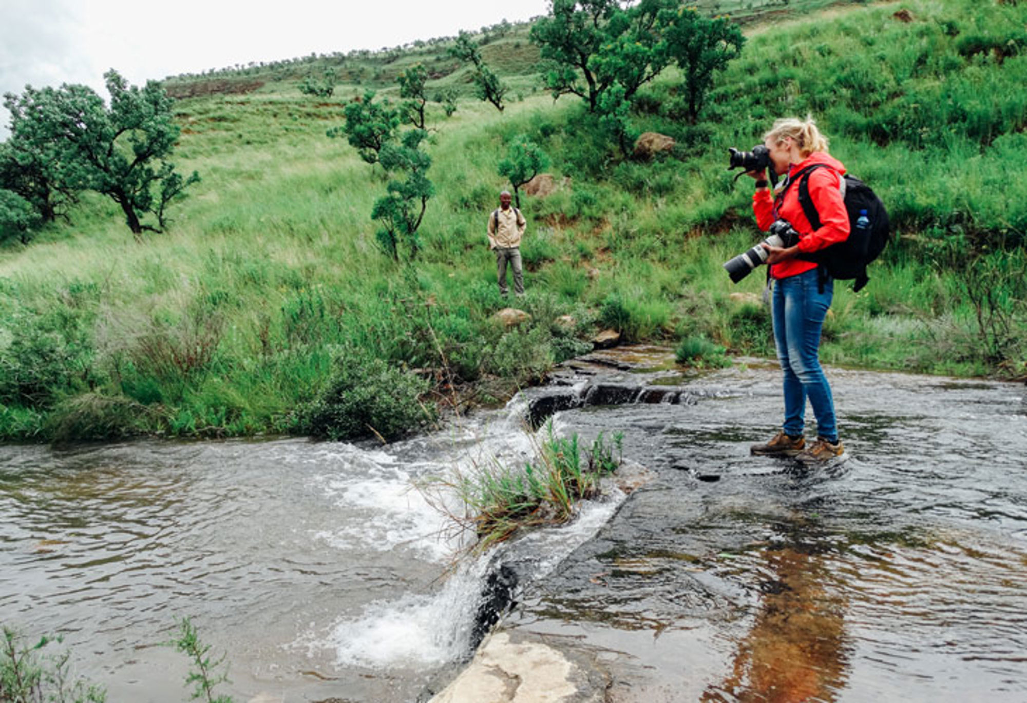 Fotograaf met camera op een rots bij een beek in groen heuvellandschap.