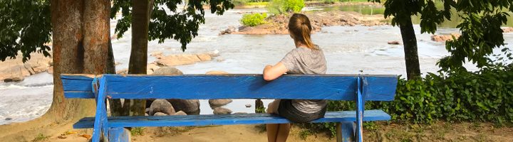 Reiziger op blauwe bank aan de rivier in de Surinaamse jungle, met rotsen en groen op de achtergrond.