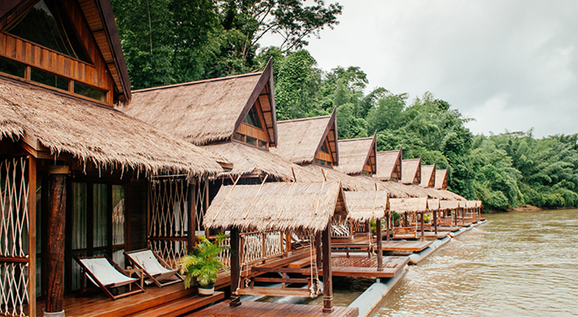 Rij drijvende bungalows met rieten daken aan een rivier, omringd door tropisch groen.