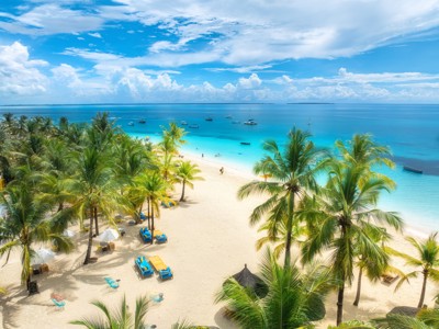Palmenstrand op Zanzibar met wit zand, ligbedden en turquoise zee met boten onder een blauwe lucht.