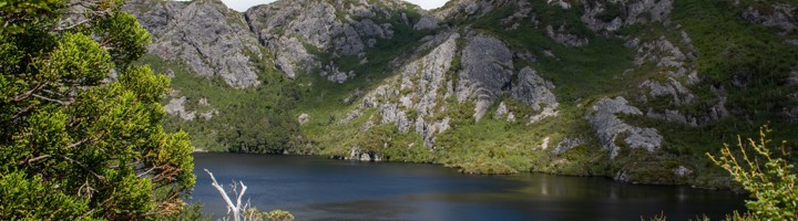 Bergmeer in Tasmanië met rotsachtige bergen en groen struikgewas onder een blauwe lucht.