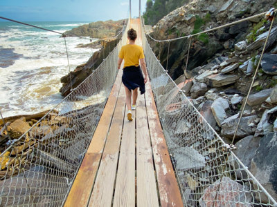 Wandelaar op hangbrug langs de ruige kust van Zuid-Afrika, tussen rotsen en de oceaan.