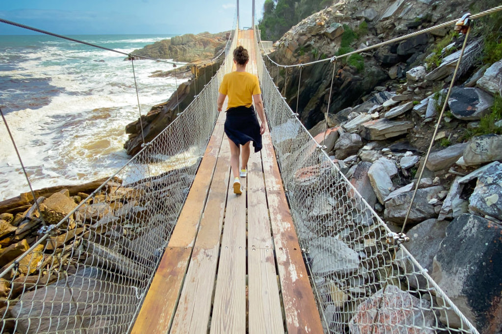 Wandelaar op hangbrug langs de ruige kust van Zuid-Afrika, tussen rotsen en de oceaan.