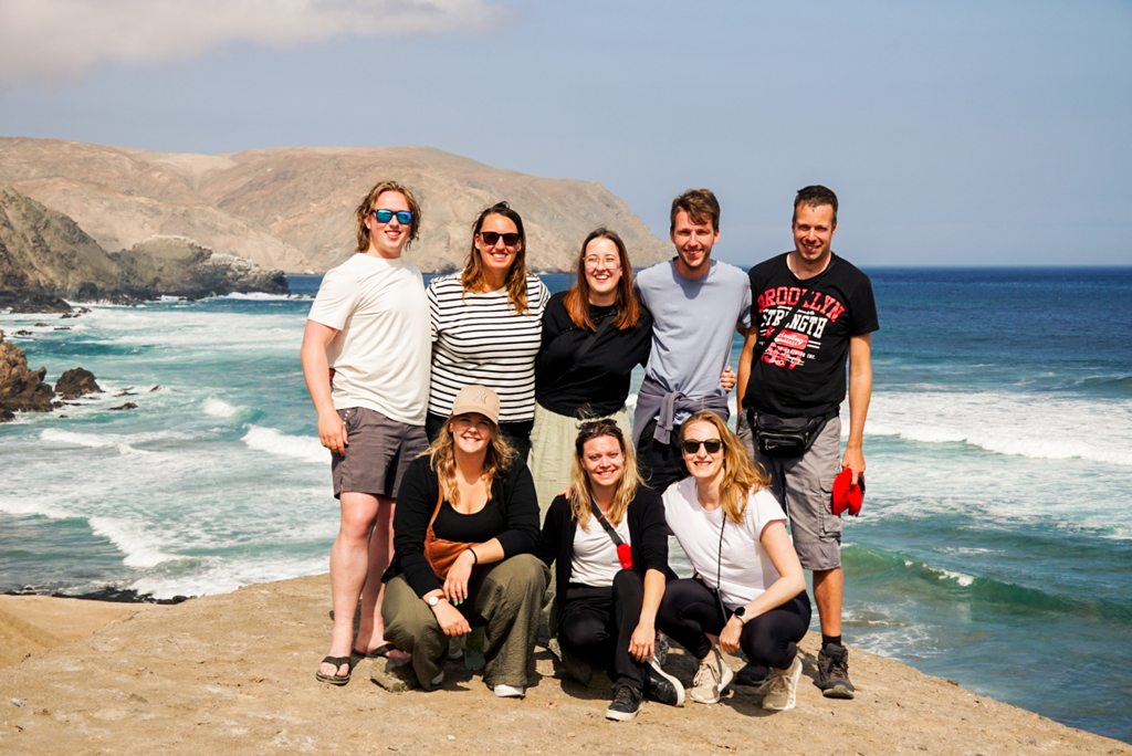 Groep 22-35ers poseert aan de kust in Peru met zee en rotsachtige heuvels op de achtergrond.