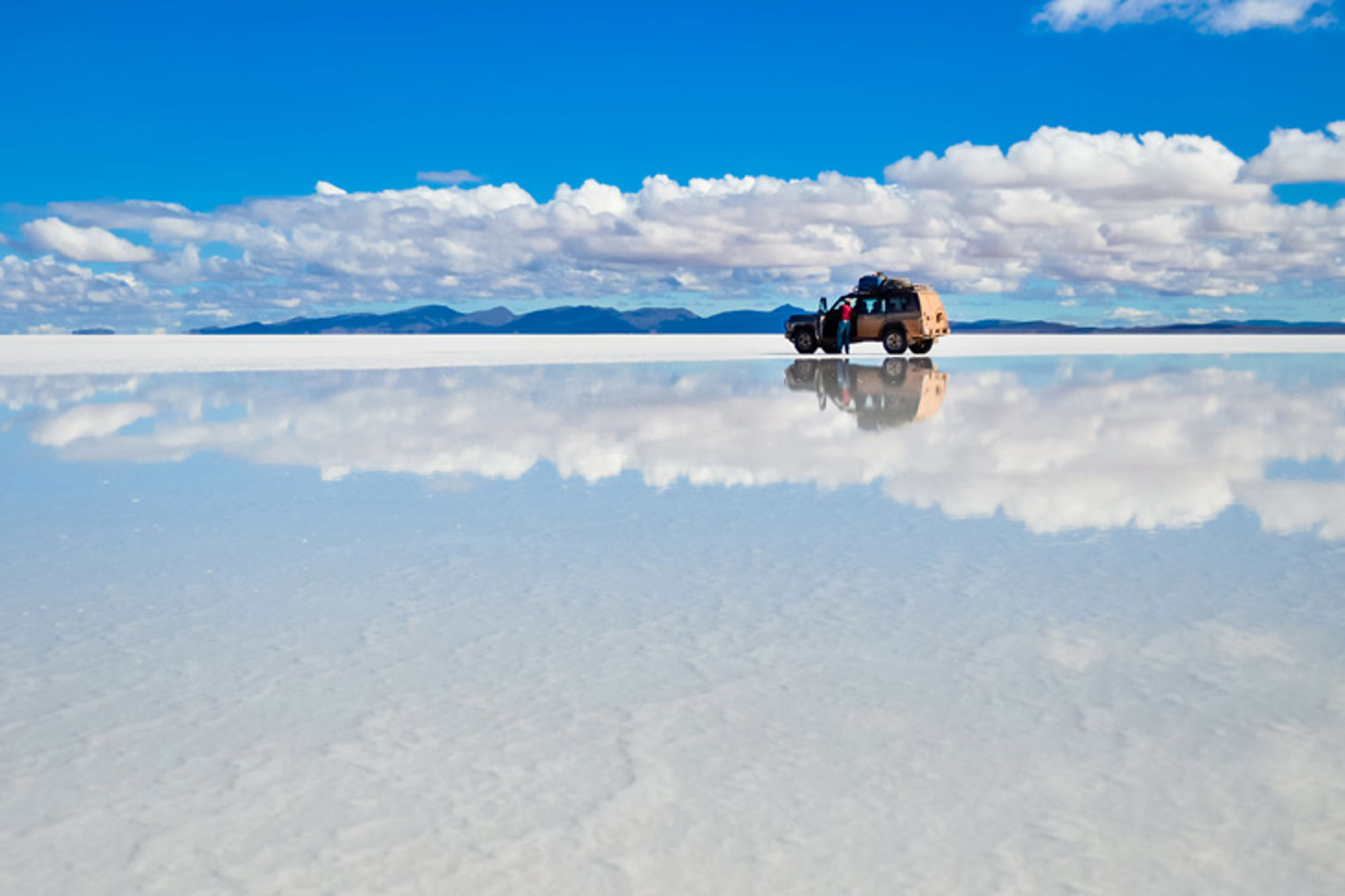 4x4 en reiziger op een spiegelende zoutvlakte met wolkenlucht en bergen op de horizon.