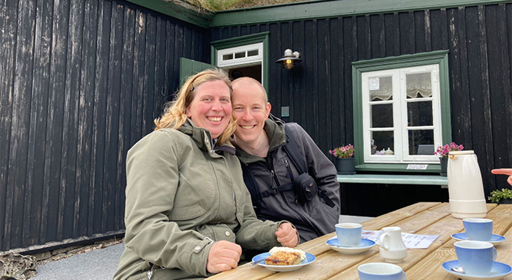 Koppel geniet van koffie en gebak aan een houten tafel bij een zwart houten huis met groene kozijnen.