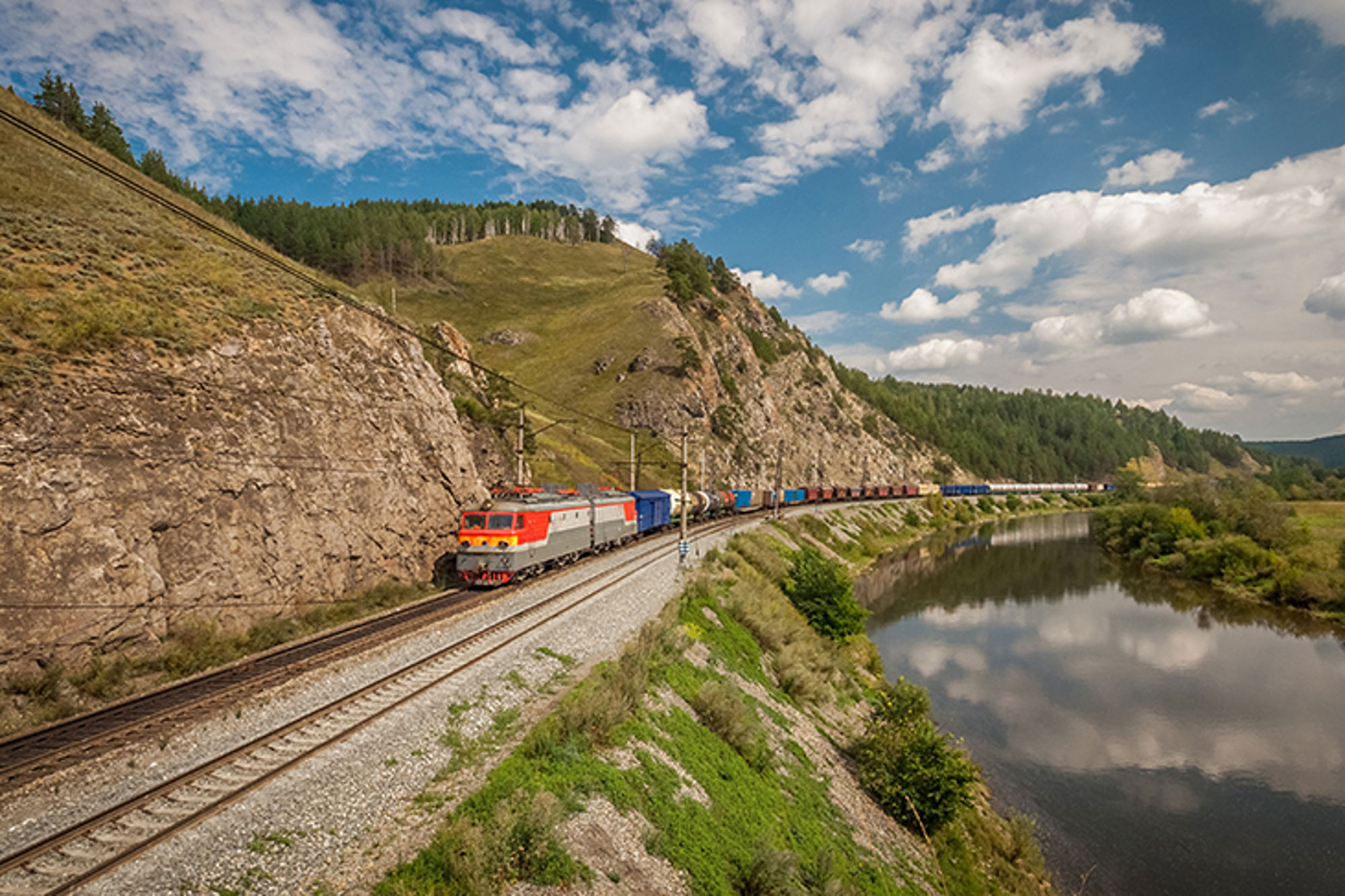 Goederentrein rijdt langs rotswand en rivier door groen heuvelachtig landschap onder blauwe lucht met wolken.